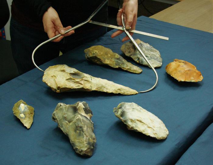 Several flint stone handaxes, some round, some pointed and of many different sizes, sit on a table. The largest, a giant handaxe, is being measured using wide spring callipers.