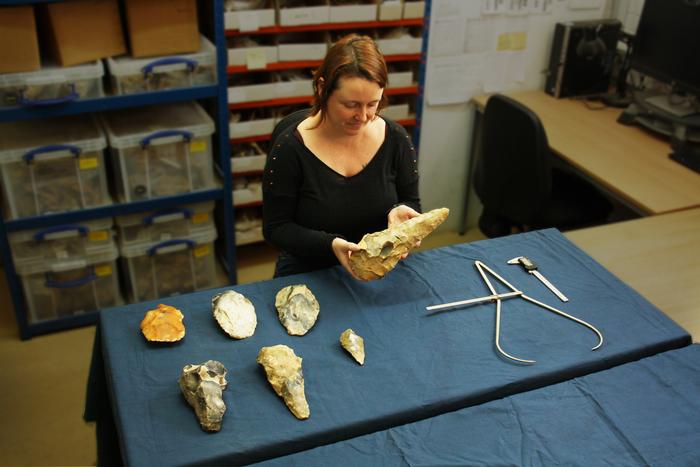 An archaeologist inspects a very large flint handaxe that she is holding in her hands. Several other flint stone handaxes, some round, some pointed and of many different sizes, sit next to her on a table, along with two types of callipers for measuring.