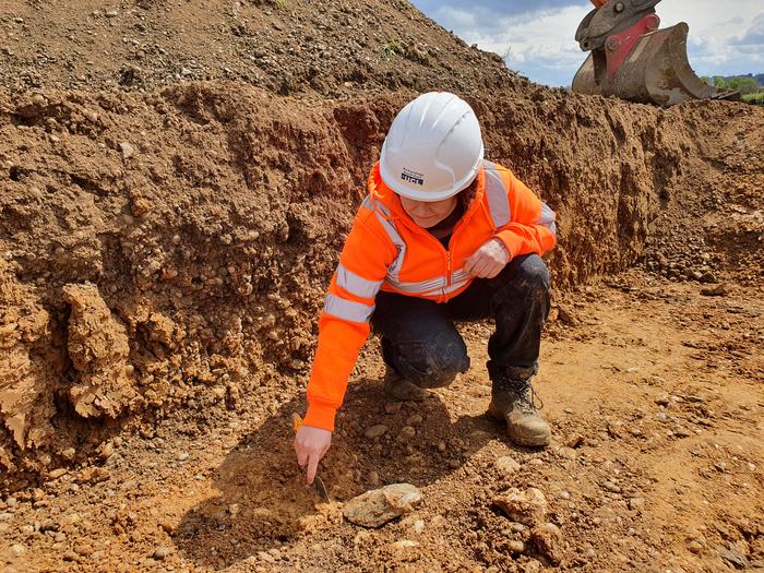 An archaeologist excavating using a trowel in a trench. They are wearing a hard hat and high visibility clothing. Just visible in the foreground next to the archaeologist’s trowel is a large flat stone, which is one of the handaxes.