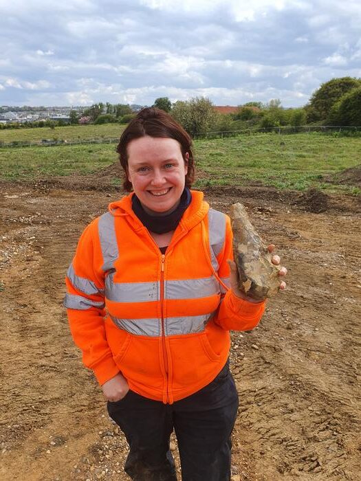 An archaeologist standing on an excavation site smiles as she holds up one of the handaxes, fresh from the ground.