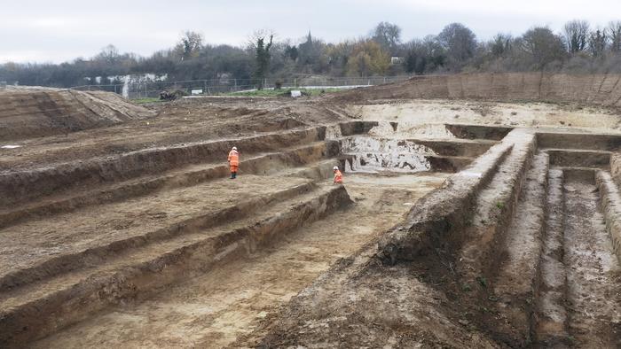 A large scale excavation of deep trenches with multiple stepped sides. Two diminutive figures of archaeologists working with hoes offer a sense of scale.